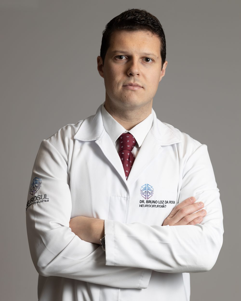 Dr. Bruno Loz, neurocirurgião, de jaleco branco com o nome bordado e braços cruzados, olhando para a câmera
